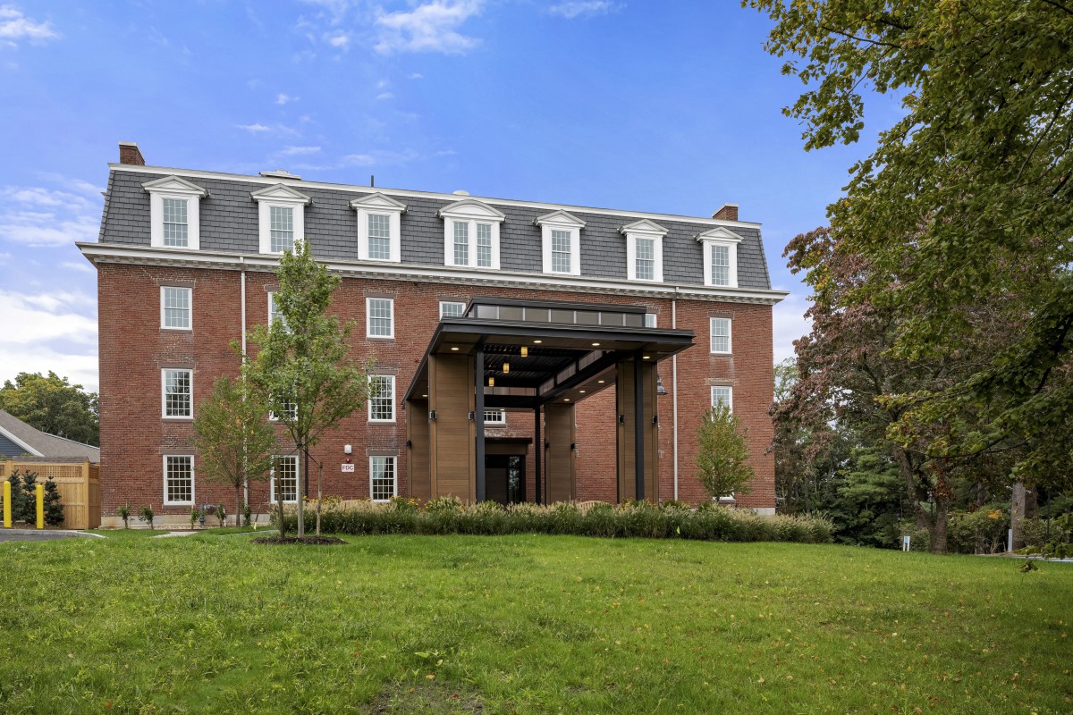 A brick building with a covered entrance, surrounded by trees and grass under a clear blue sky.