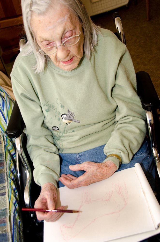 An elderly woman in a wheelchair sketching on a piece of paper.