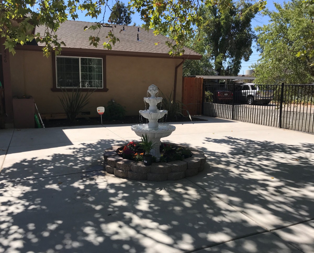 A sunny patio with a decorative fountain and surrounding trees casting shadows.
