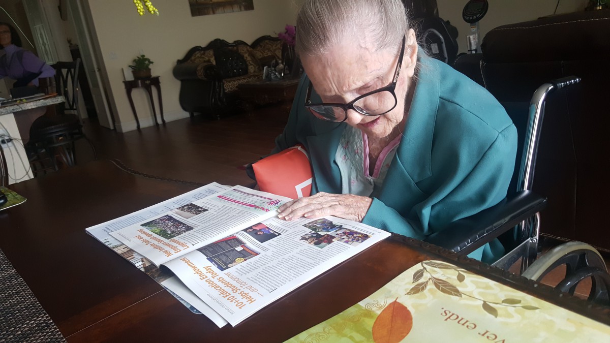 An elderly woman wearing glasses reads a magazine at a table indoors.