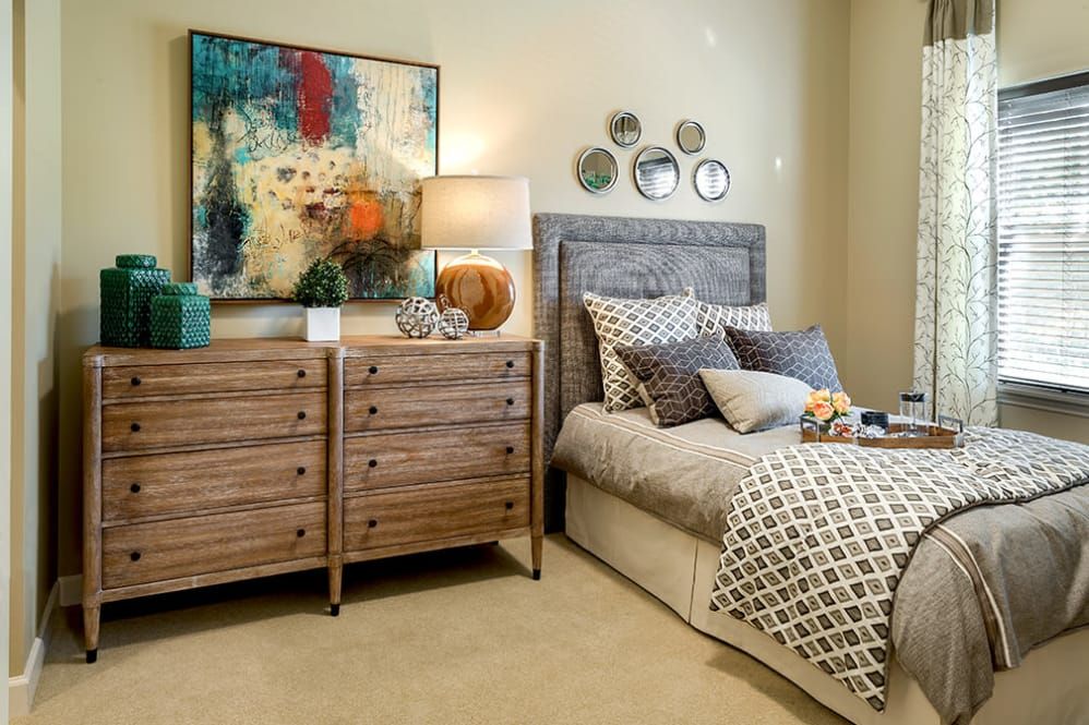A modern bedroom with elegant furnishings, featuring a textured gray headboard, wooden dresser, abstract artwork, and a cozy lamp.