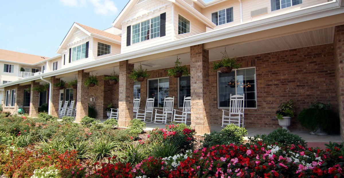 A charming building with a porch featuring rocking chairs, surrounded by colorful flowers and greenery.