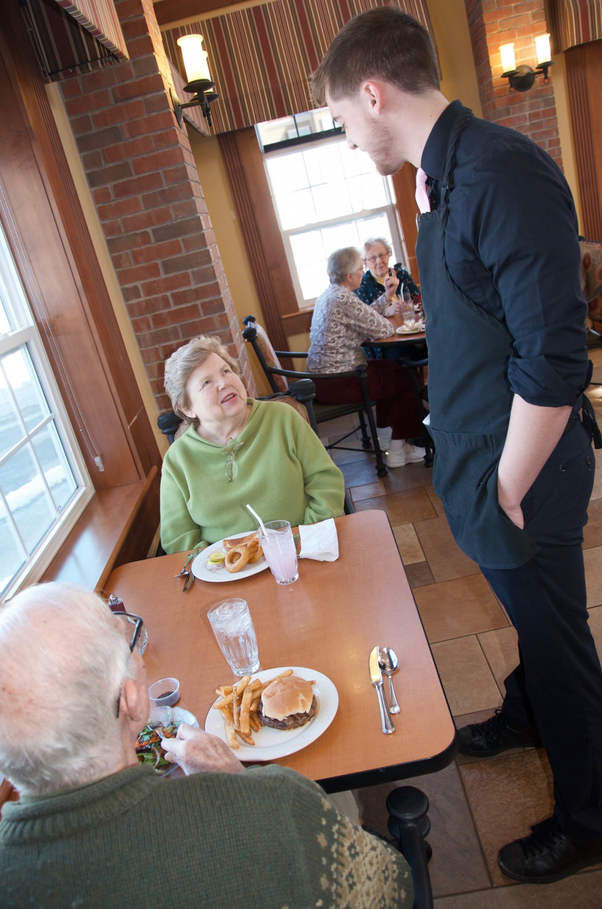 An elderly couple seated at a restaurant table, engaging in conversation with a waiter.
