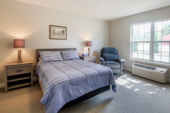 A cozy bedroom with a neatly made bed, a lamp on a nightstand, and a comfortable chair by the window.