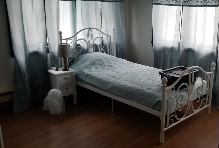 A cozy bedroom featuring a single bed with wrought iron frame, a nightstand with a lamp, and soft curtains lining the windows.