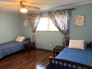 A cozy bedroom with two blue-covered beds, wooden flooring, and a ceiling fan.