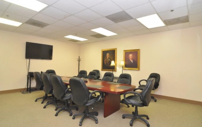 A spacious conference room with a large meeting table surrounded by office chairs, featuring portraits on the wall and a television.