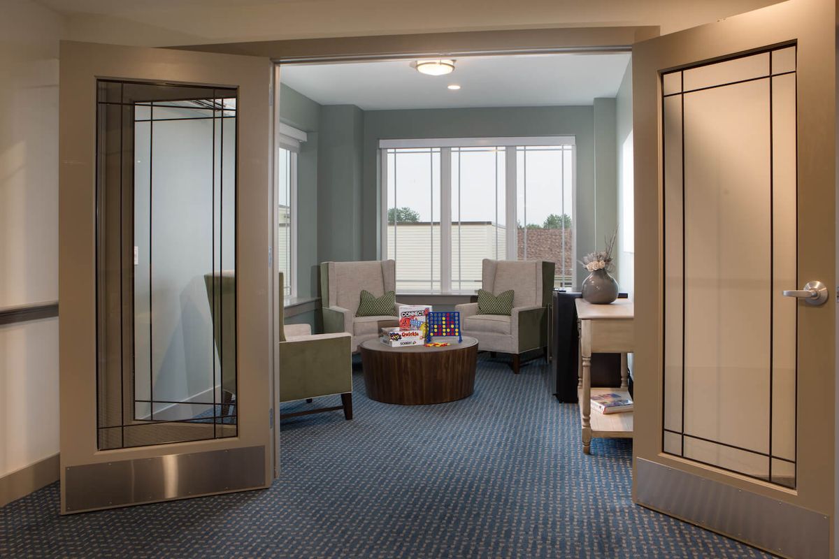 A modern waiting room with comfortable chairs and a central table, illuminated by natural light through large windows.
