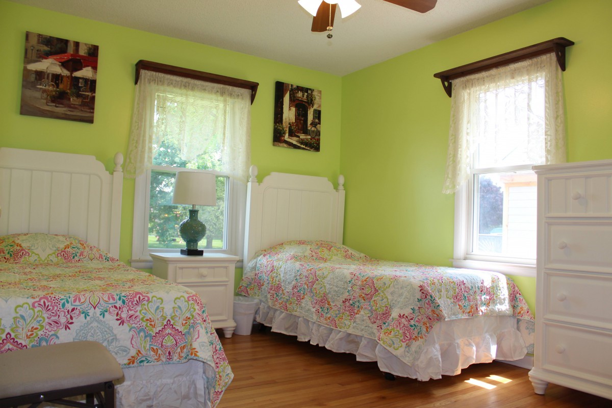 A cozy bedroom with twin beds and green walls, illuminated by natural light from two windows.