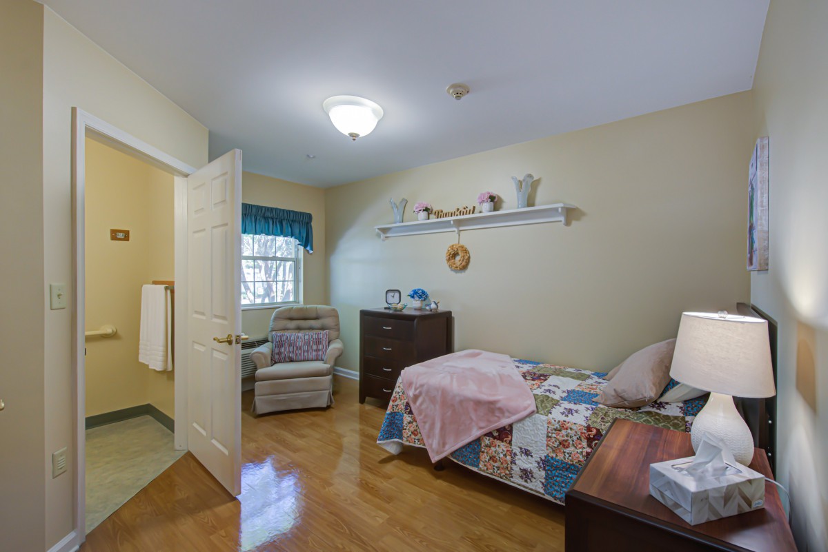 A cozy bedroom with a single bed, armchair, dresser, and decorative items on a shelf.