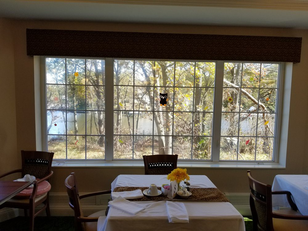 A restaurant interior with tables and chairs, featuring large windows overlooking a scenic lake and trees.