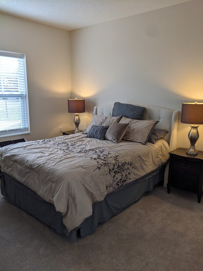A cozy bedroom with a well-made bed, flanked by two nightstands and lamps, illuminated softly by natural light from a nearby window.
