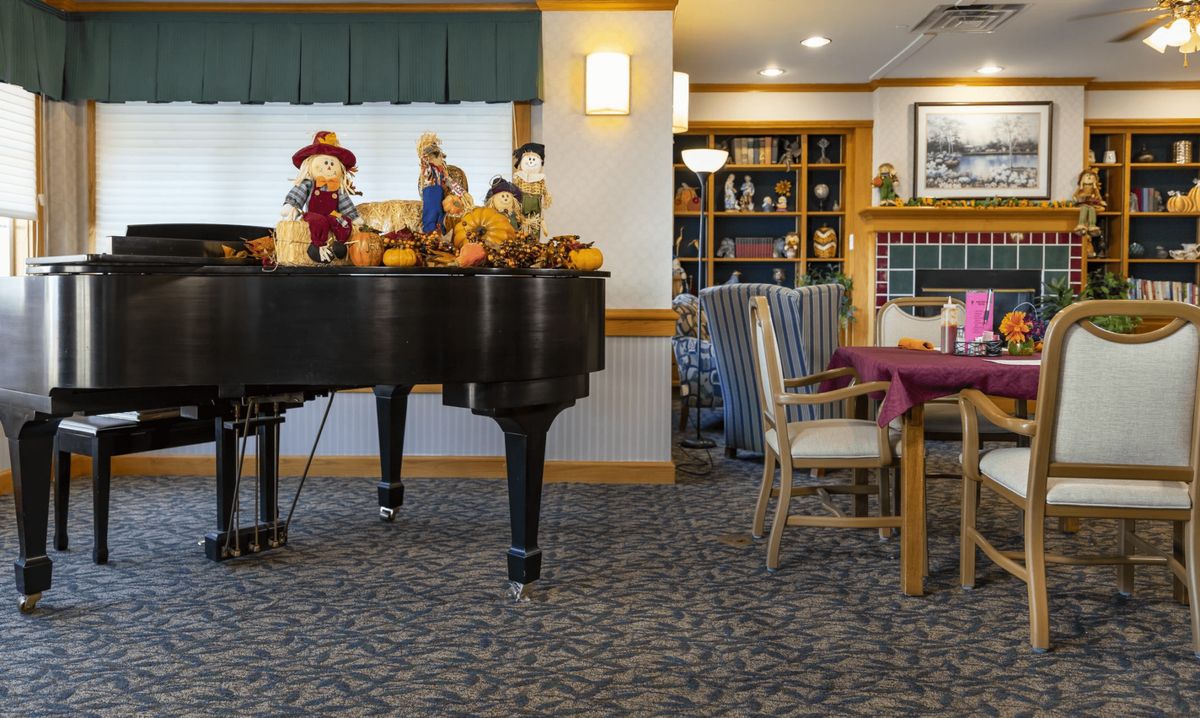 A cozy living room with a grand piano decorated with scarecrows and pumpkins, surrounded by comfortable seating and bookshelves.