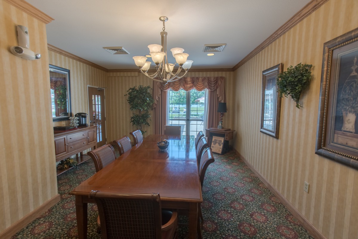 A dining room with a long table and chairs, illuminated by a chandelier, featuring large windows with curtains and decorated with artwork and plants.