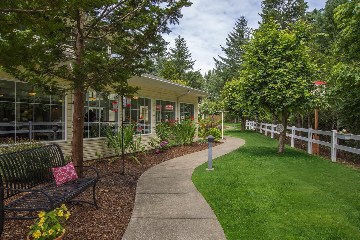 A scenic garden pathway lined with lush greenery and trees, bordered by a white fence.