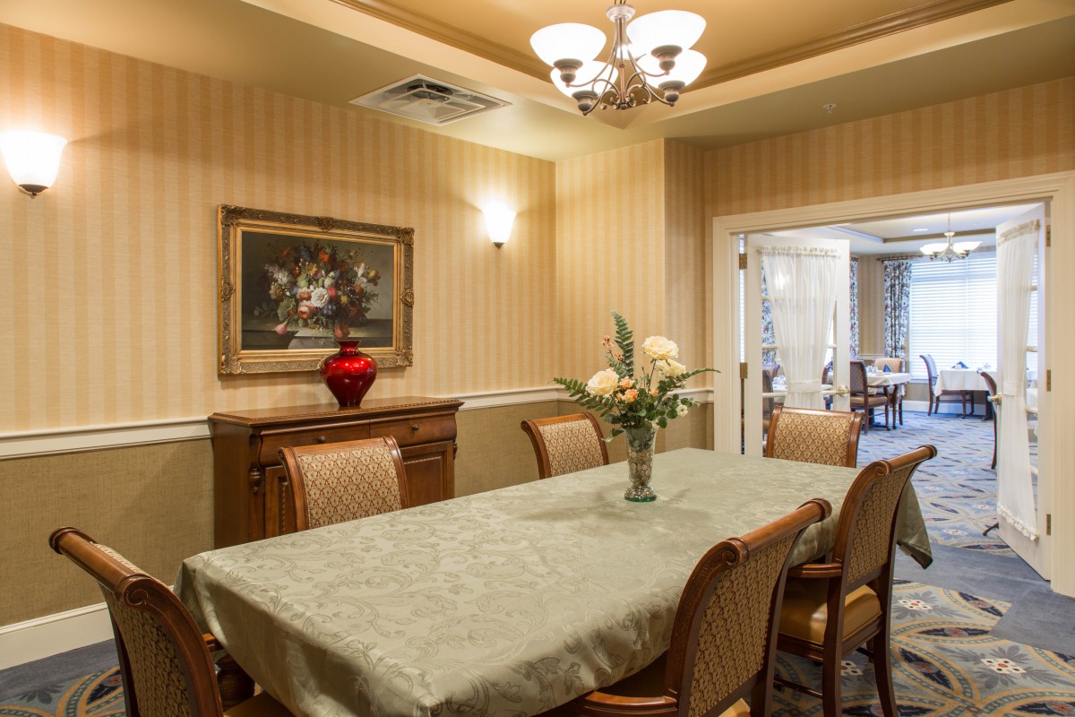 A warmly decorated dining room with a long table and upholstered chairs, adorned by a chandelier and a painting on the wall.
