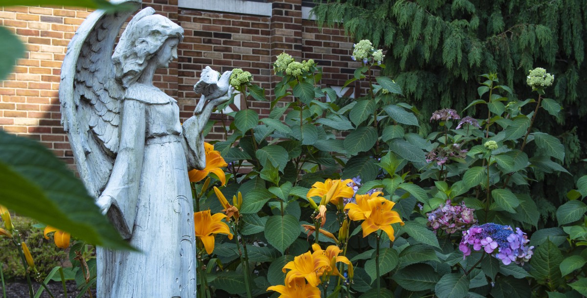 A garden with a statue surrounded by vibrant yellow flowers and lush greenery against a brick wall backdrop.
