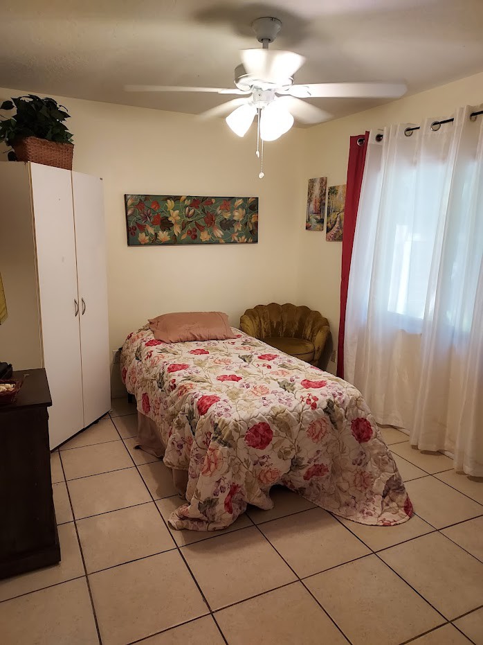 A cozy bedroom with a single bed adorned with a floral bedspread, featuring a ceiling fan and natural light through the curtains.