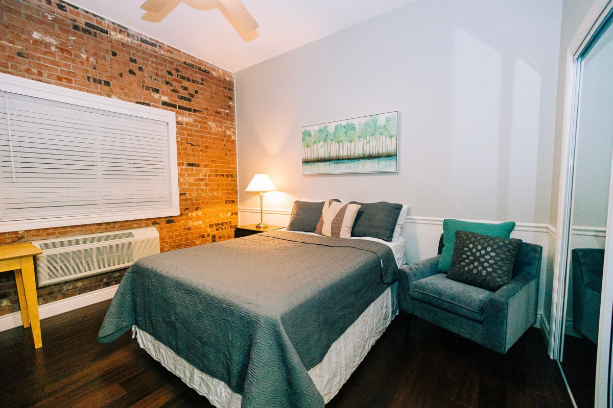 A cozy bedroom with a brick accent wall, featuring a bed with teal bedding and a soft armchair beside a lamp.
