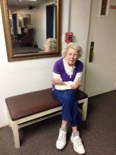 An elderly woman sitting on a bench indoors, with a mirror behind her.