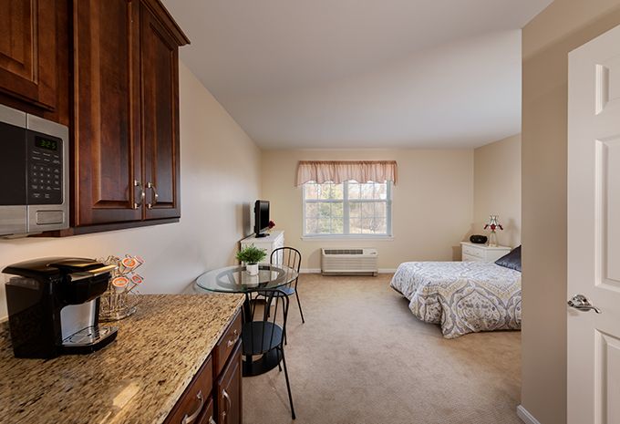 A cozy bedroom interior featuring a bed, table with chairs, cabinetry, and a window providing natural light.