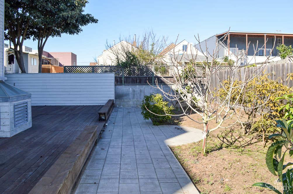 A sunny garden with a paved pathway, trees, and a wooden deck.
