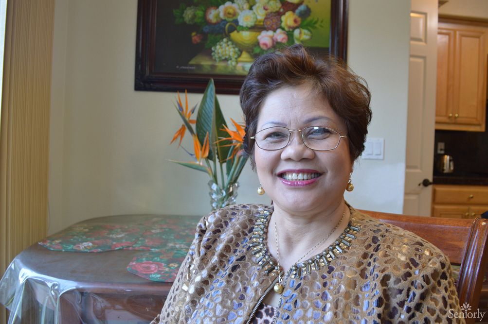 A woman with glasses smiling indoors, sitting near a table with flowers and a painting on the wall.