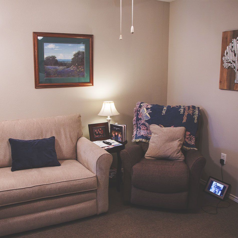 A cozy living room with a couch and armchair, decorated with paintings and illuminated by a small lamp.