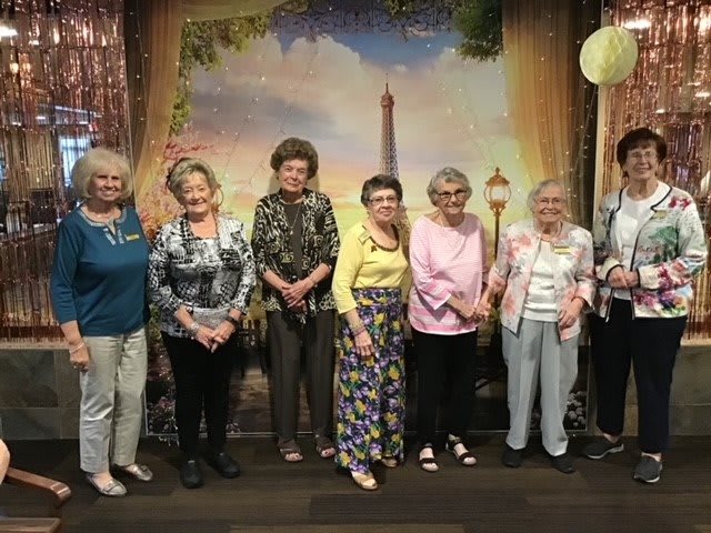 A group of elderly women posing together in front of a scenic backdrop, all smiling and dressed in colorful clothing.