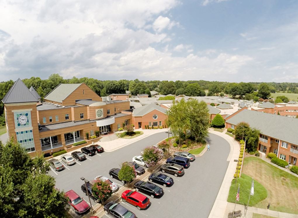 An aerial view of a suburban area with a building and parking lot surrounded by greenery under a cloudy sky.