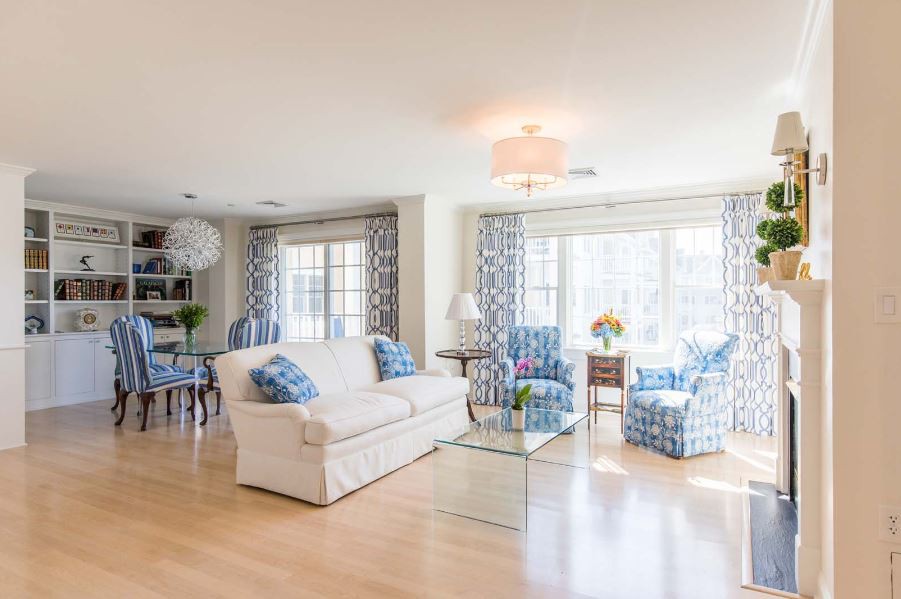 A bright and spacious living room with a white sofa, patterned blue armchairs, and large windows letting in natural light.