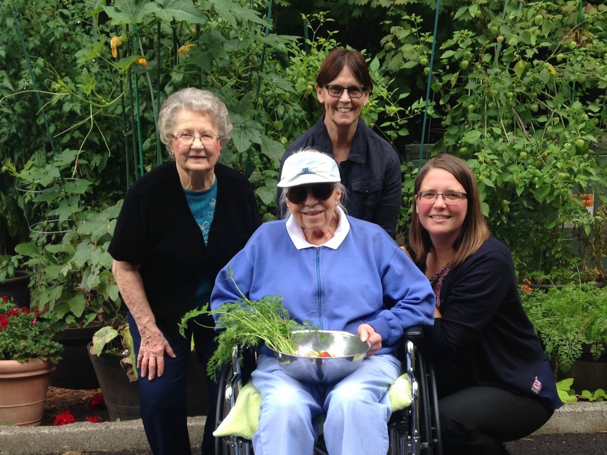 A group of people smiling outdoors in a garden, with one person in a wheelchair holding plants.