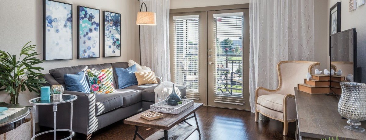A cozy living room with a modern gray sofa, decorative cushions, and a stylish coffee table in front of French doors with blinds.