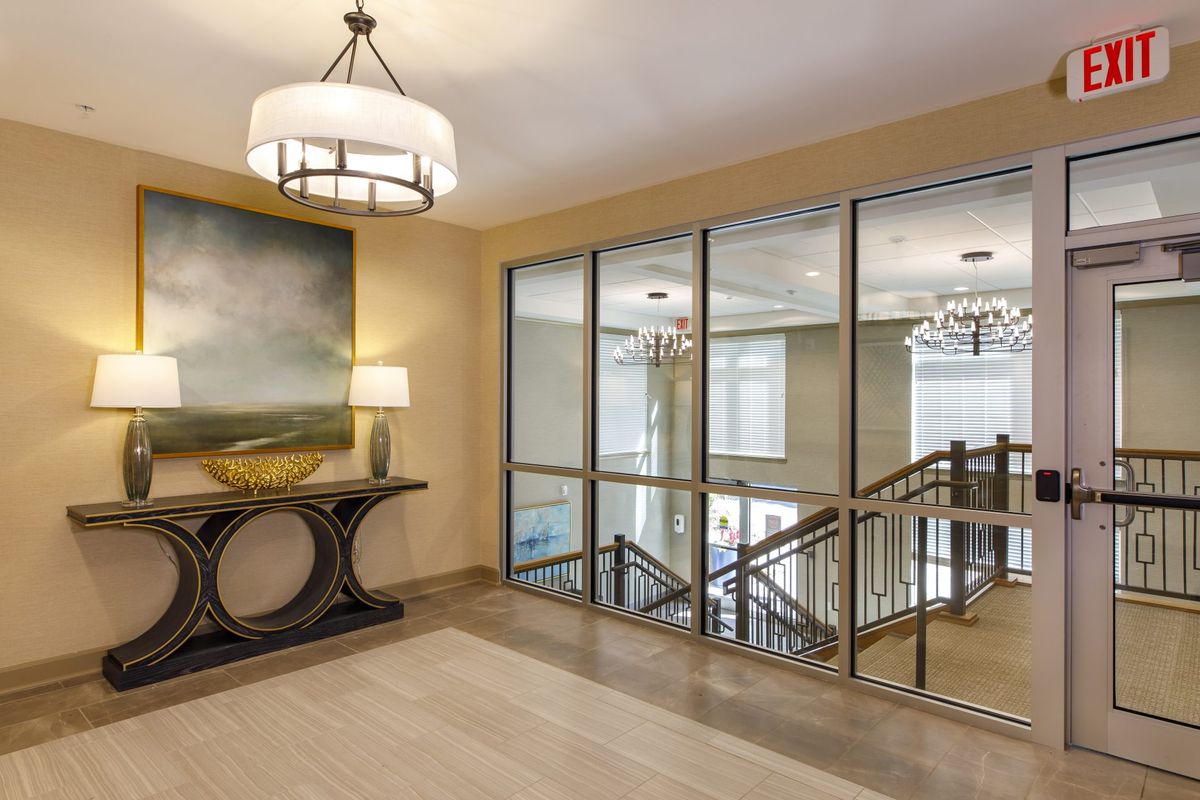 A modern lobby featuring a console table with artwork, a glass partition, and a staircase with chandeliers.