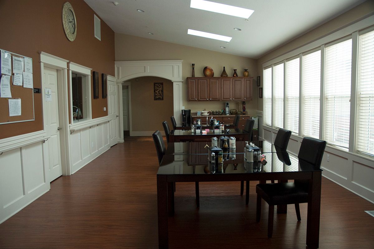 A spacious dining room with wooden flooring, featuring a large table surrounded by chairs, and a kitchen area in the background.