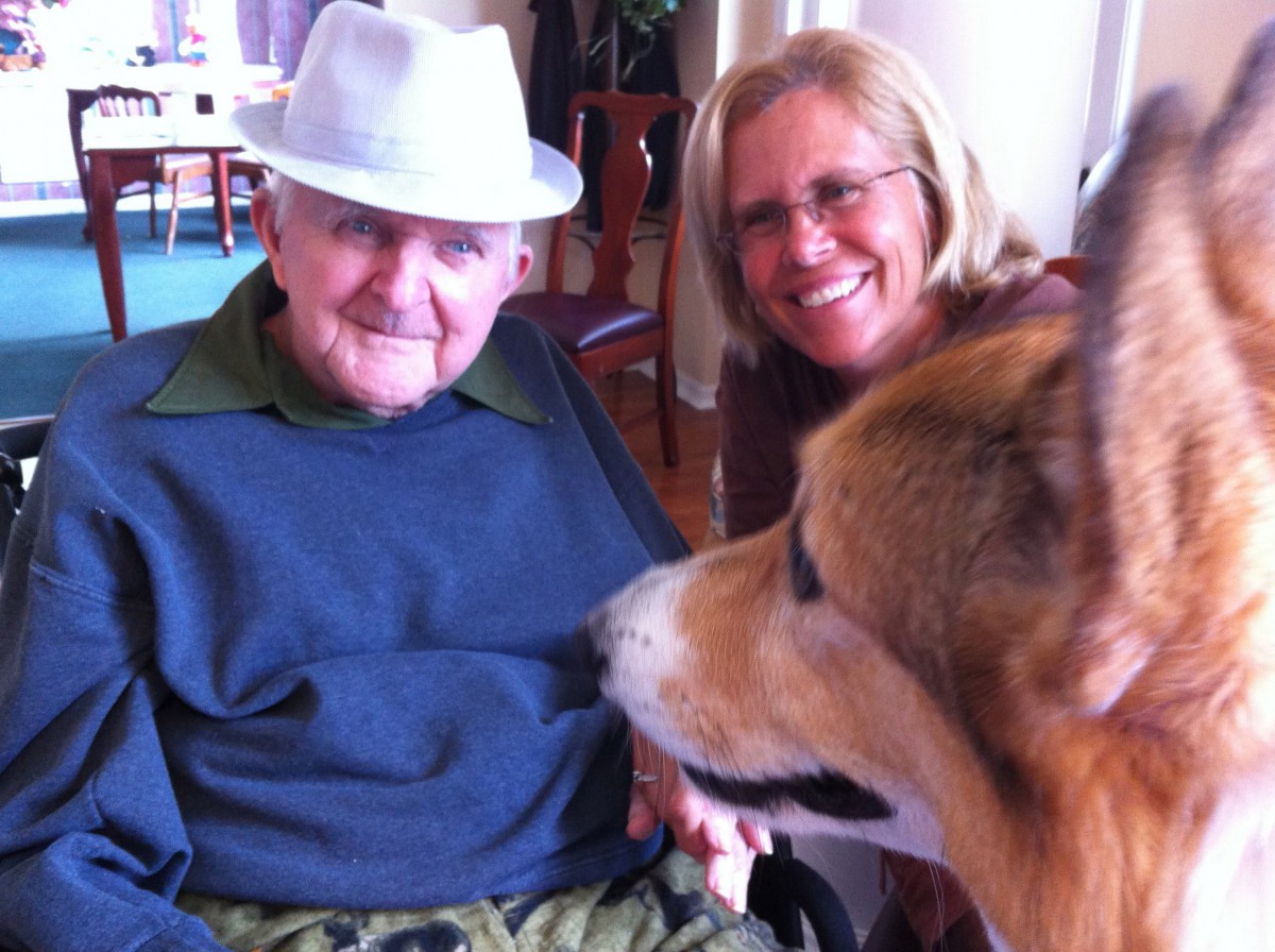 An elderly man wearing a hat sits next to a smiling woman and a dog indoors.