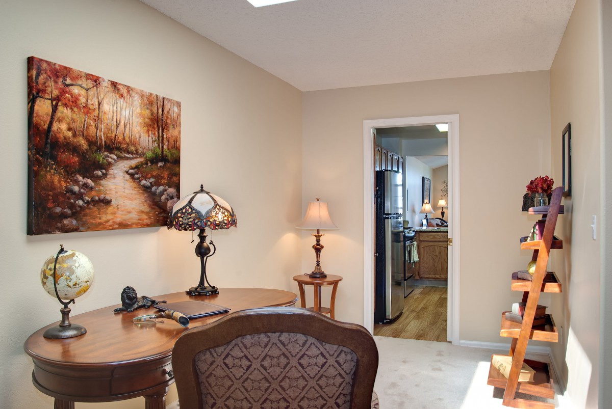 A cozy home office featuring a desk, lamps, and artwork on the wall with a view into the kitchen.