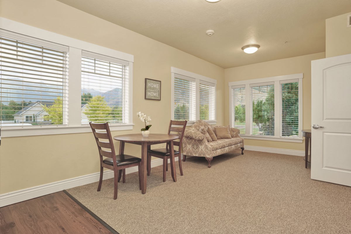 A cozy living room with a small dining table, chairs, and a sofa, surrounded by large windows.