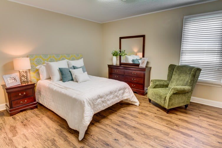 A cozy bedroom with a comfortable bed, green armchair, dresser, and wooden flooring.
