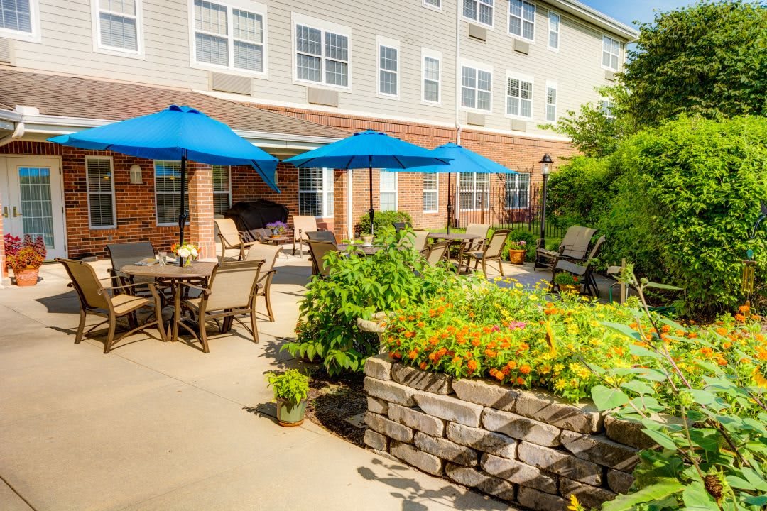 A sunny patio with tables and blue umbrellas, surrounded by a lush garden.