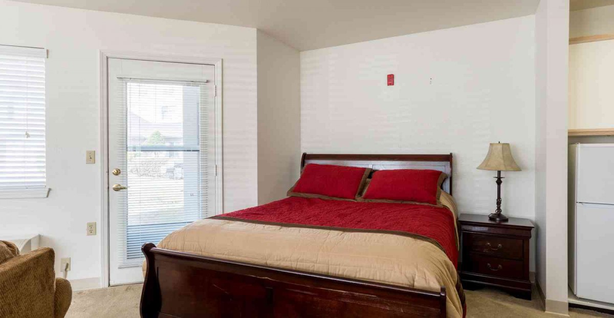A neatly made bed with red and beige bedding in a bright bedroom.
