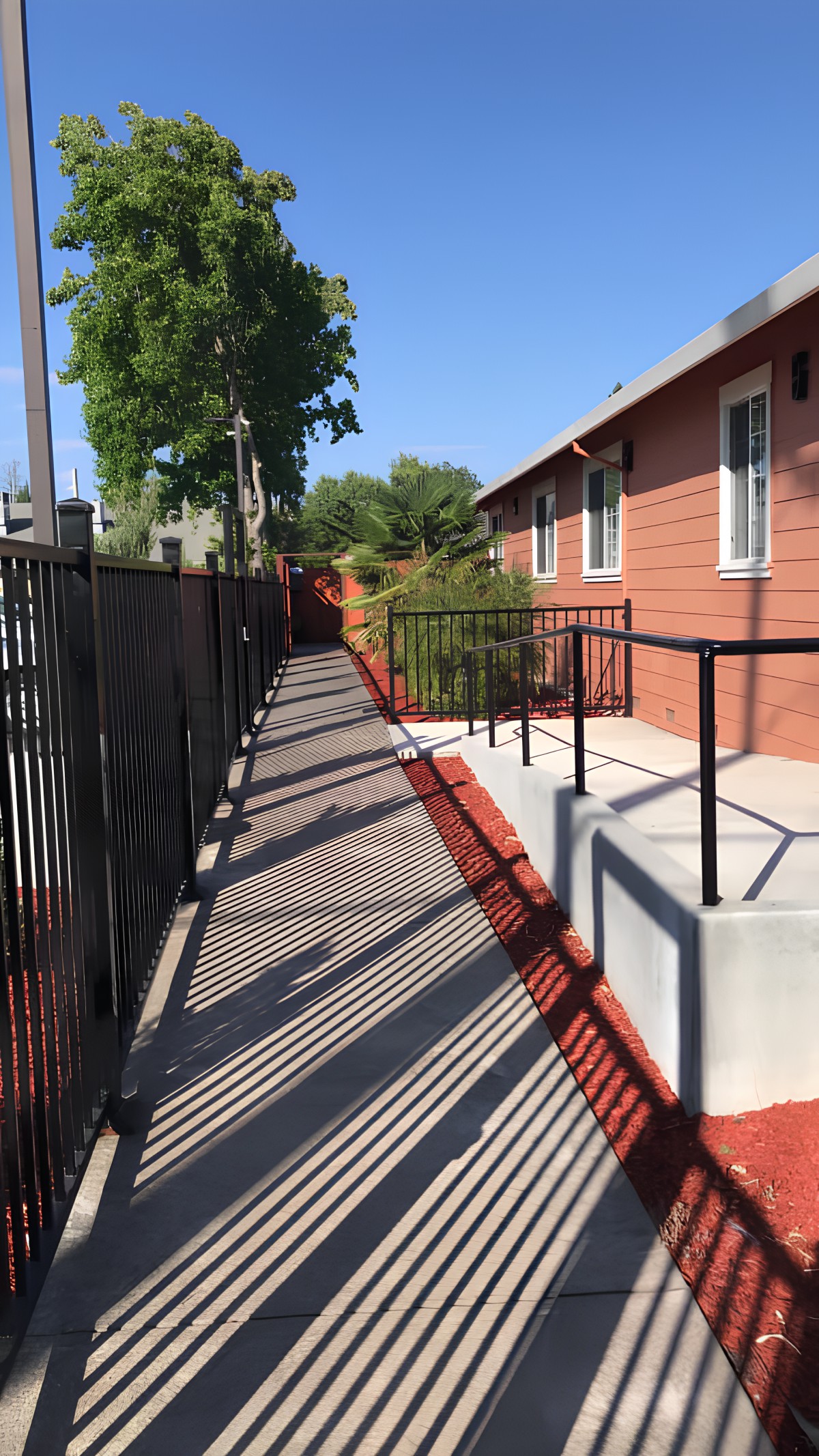 An outdoor ramp alongside a building with a black metal fence and shadows cast on the pathway.