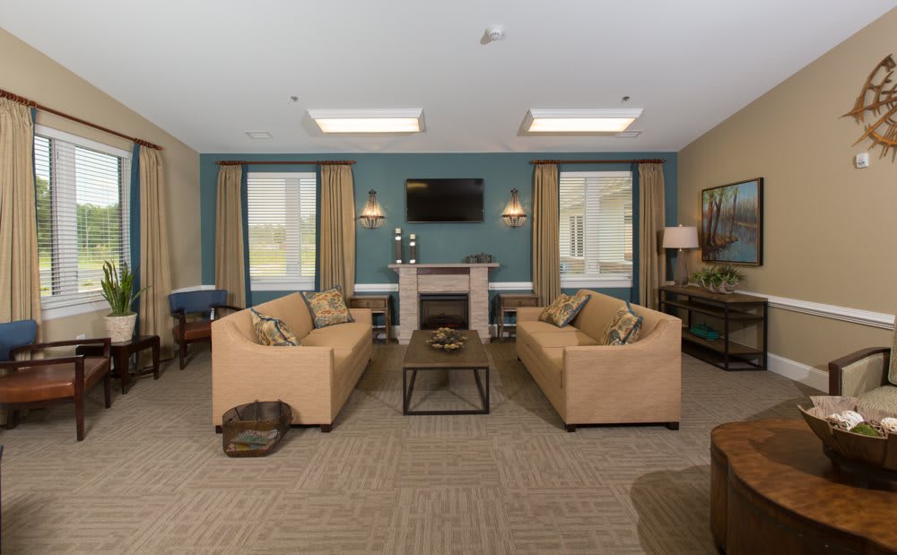 A cozy living room featuring two beige sofas, a wooden coffee table, and a fireplace with a mounted television above it. The room is brightly lit by natural light coming through the windows with blue curtains.