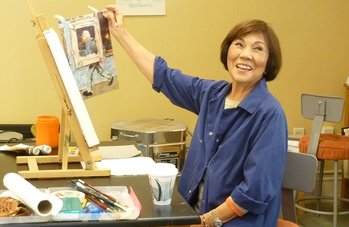 A smiling artist holds up her painting while working at an easel.