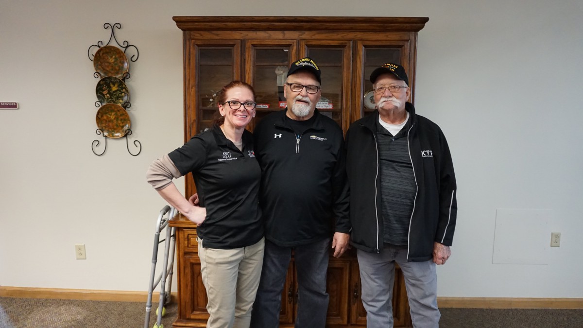 A group of three people standing together indoors, smiling at the camera.
