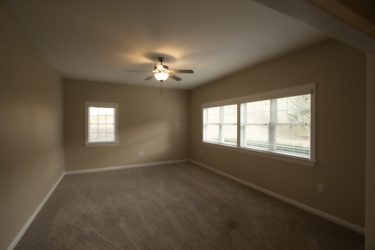 An empty room with carpet flooring, several windows, and a ceiling fan.