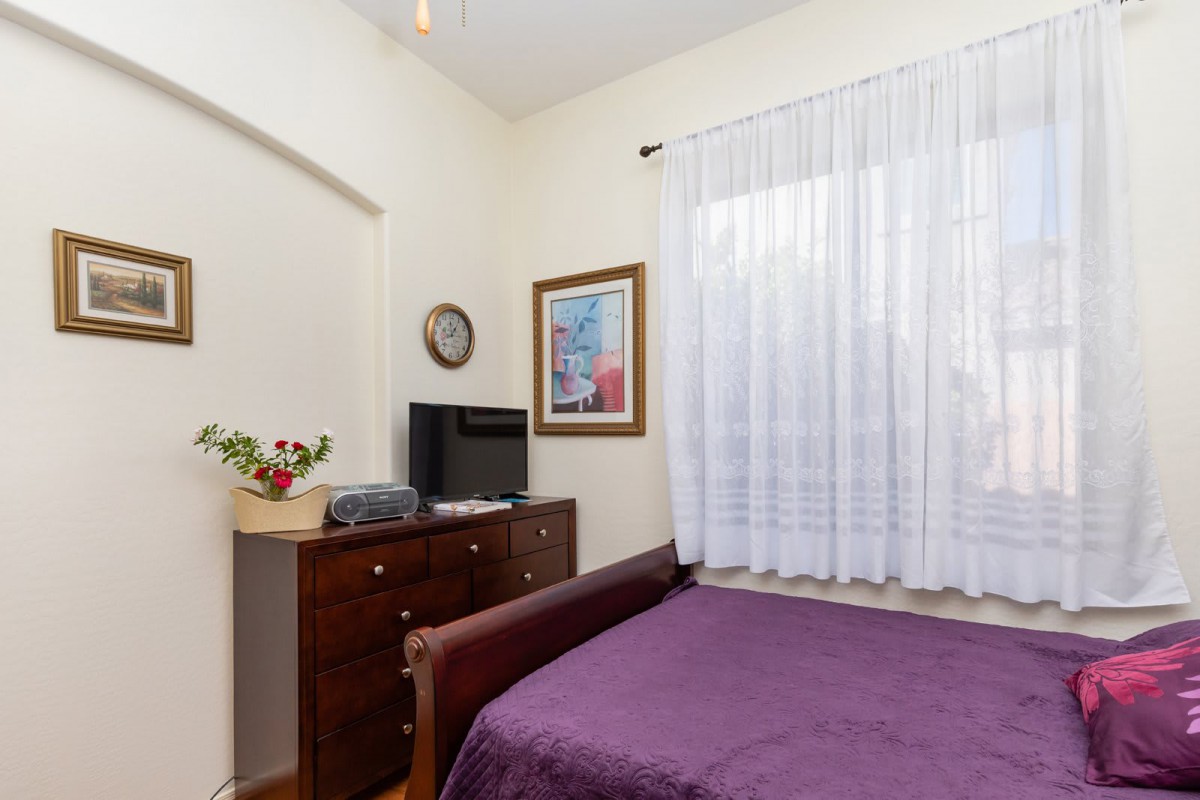 A cozy bedroom with a purple bedspread, a dresser with a TV, and a window with sheer curtains.