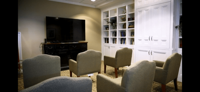 A cozy living room featuring a large television, several armchairs, and built-in bookshelves.