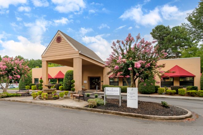 A building entrance with landscaping and signage, featuring a parking area and flowering trees.