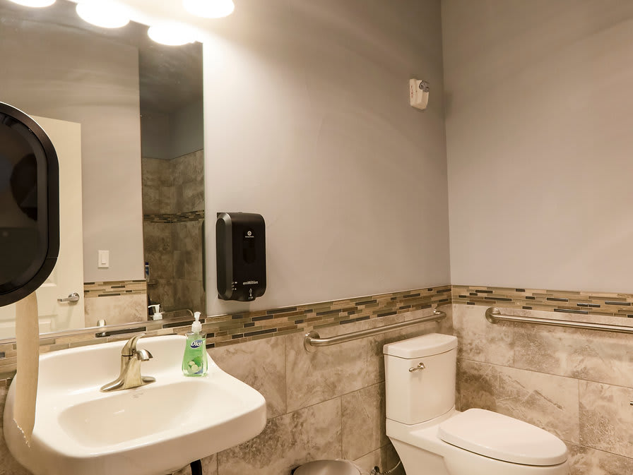 A well-lit bathroom with a sink, toilet, and mirror featuring a soap dispenser.